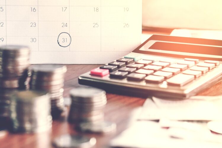 A close-up on a desk with stacks of coins, documents, a calculator, and a calendar with a date circled on it.