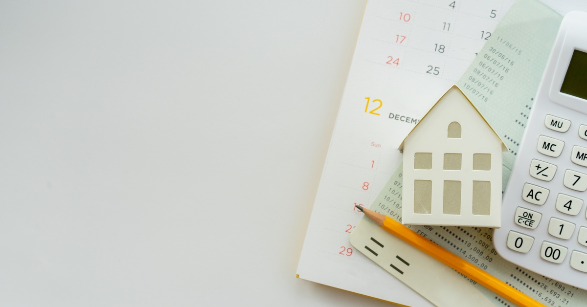 A close-up of a calendar with a small, model home placed on top of it, documents, and a calculator.