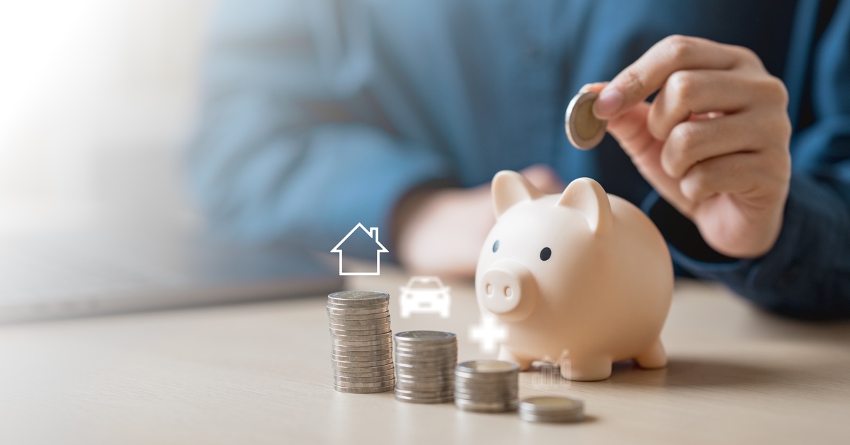A close-up of a person wearing blue placing a coin into a piggy bank next to a stack of coins.