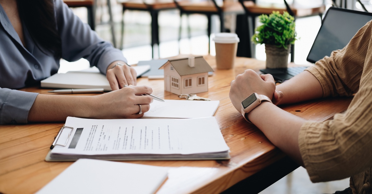 Two people sitting across from each other at a table with a model home, computer, and documents on the table.