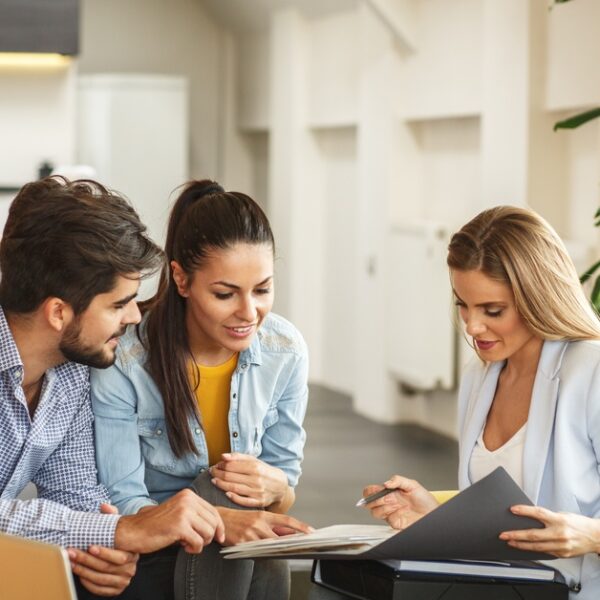 A couple sitting together across from a real estate agent flipping through a portfolio of documents in an office space.