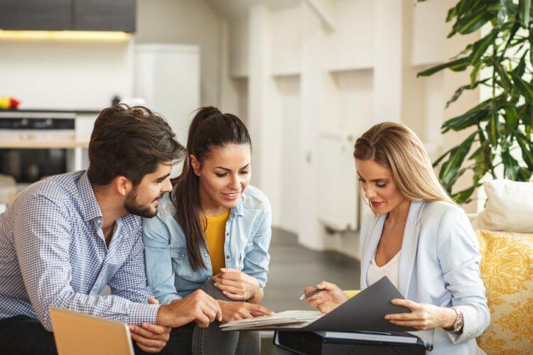 A couple sitting together across from a real estate agent flipping through a portfolio of documents in an office space.
