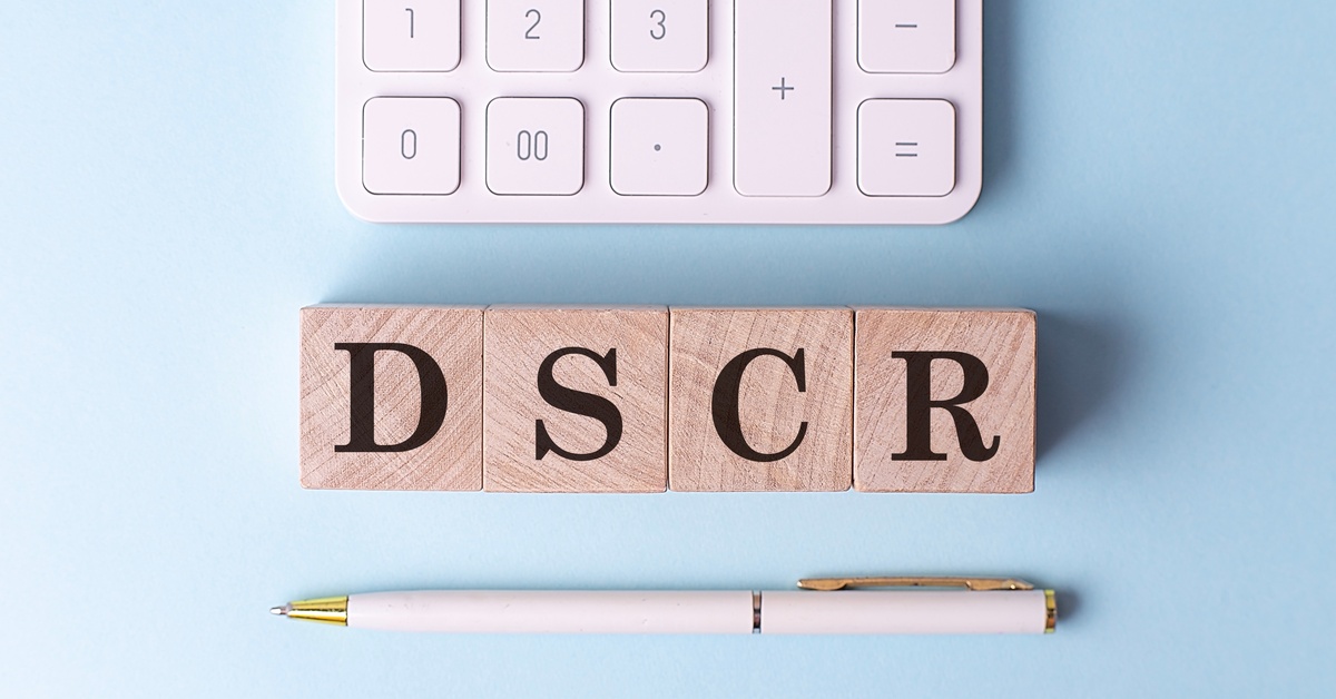 A close-up of the bottom half of a calculator, a ballpoint pen, and wooden blocks spelling out "DSCR" on a blue desk.