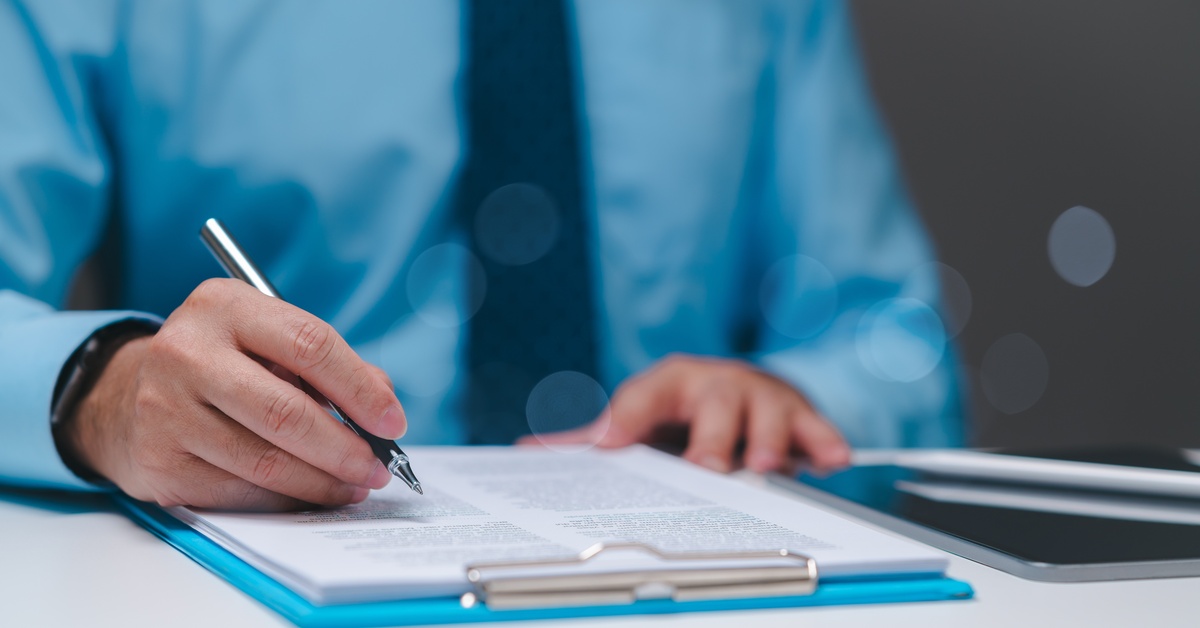 A man in a blue dress shirt sitting at a desk with a pen in hand, filling out paperwork on a clipboard.