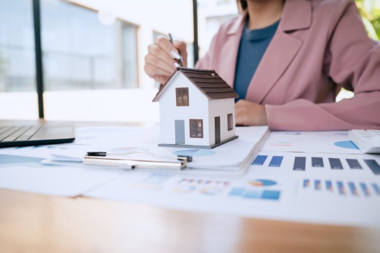 a woman reviewing financial charts at a desk beside a small house model, calculator, and printed documents.