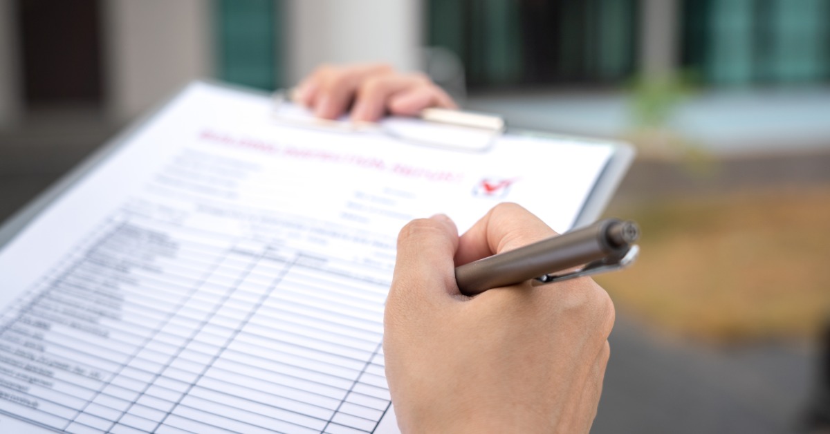 a hand holding a pen over a building inspection form, with a small house model and calculator on a desk.