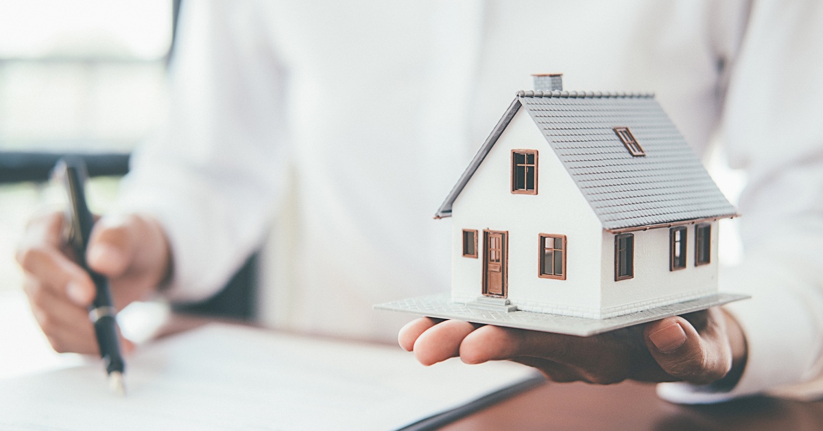 a person wearing a white shirt holds a small, white model home in one hand and signs papers with the other.