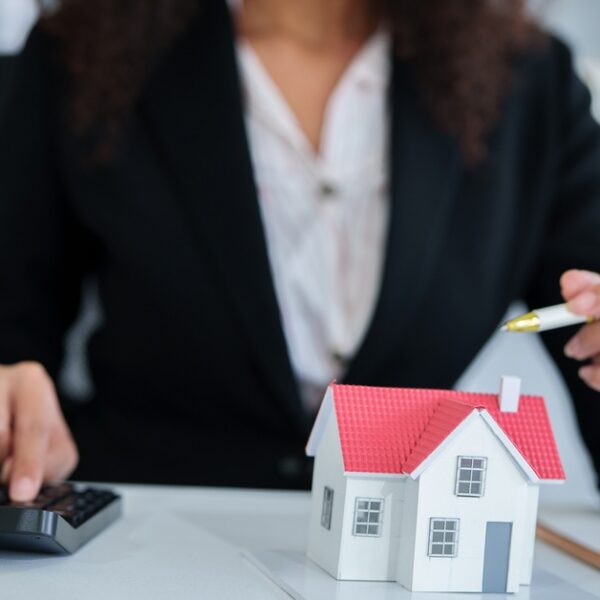 A woman in a black suit sits at a desk with a pen in one hand and the other tapping on a black calculator.