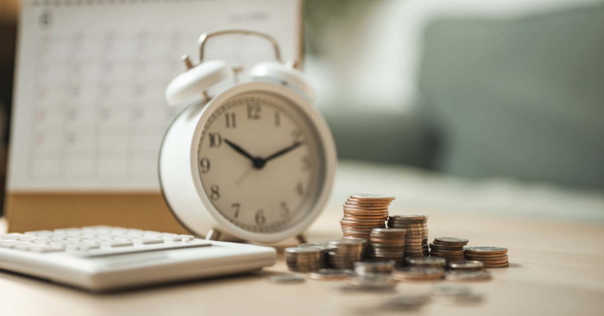 an alarm clock, calculator, calendar, and stacked coins arranged on a desk with papers and glasses.