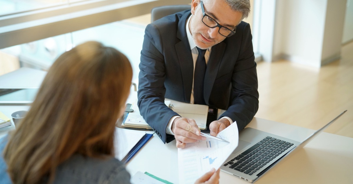 a man in glasses sitting at a desk with a laptop and papers while a woman holds a document across from him.
