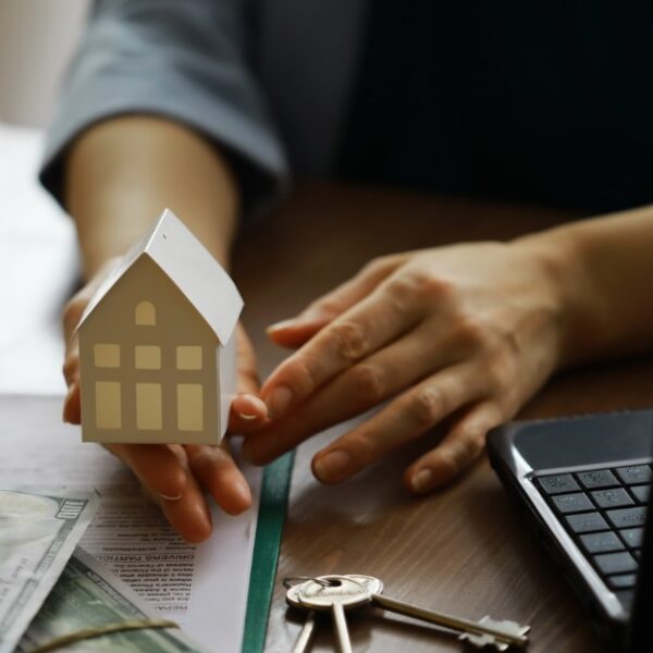 a hand holding a small house model beside cash, keys, documents, and a laptop on a wooden office table.