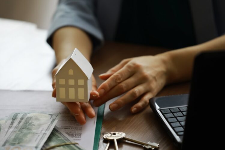 a hand holding a small house model beside cash, keys, documents, and a laptop on a wooden office table.