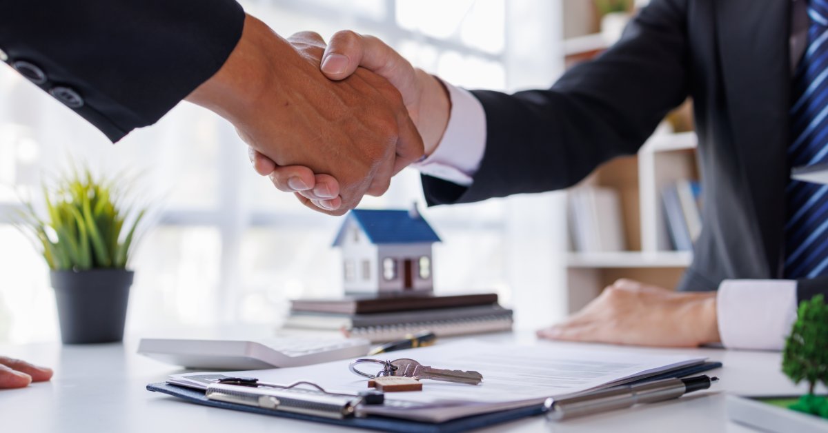 Two people shaking hands across a desk with keys, papers, a pen, a calculator, and a small house model.