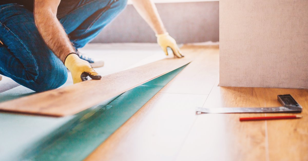A person wearing gloves laying a wood flooring plank, with a pencil and square tool on the floor nearby.