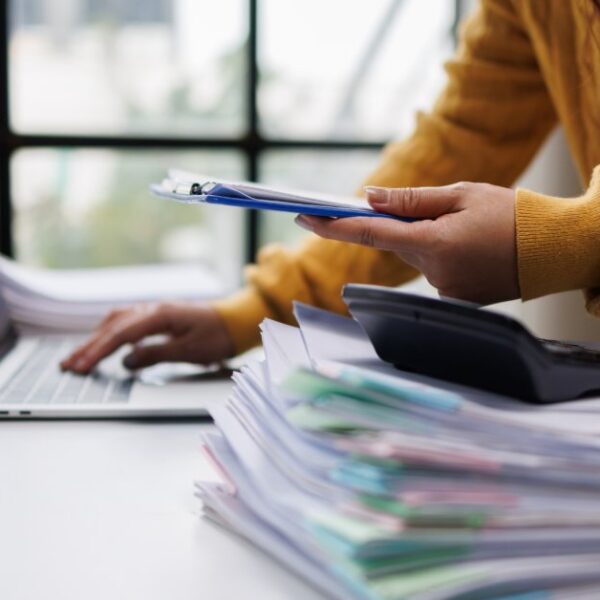 A person in a mustard sweater using a laptop beside stacked folders, a calculator, a plant, and a clipboard.