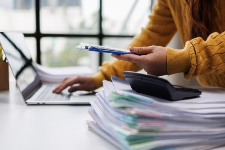 A person in a mustard sweater using a laptop beside stacked folders, a calculator, a plant, and a clipboard.