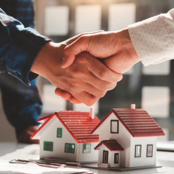 A close-up of two people shaking hands above a few model houses on a desk, with documents and a laptop nearby.