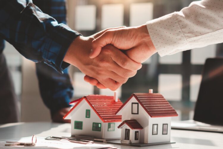 A close-up of two people shaking hands above a few model houses on a desk, with documents and a laptop nearby.