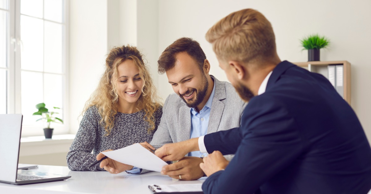 A smiling couple reviewing documents with a formally dressed man who is pointing to paperwork across an office desk.