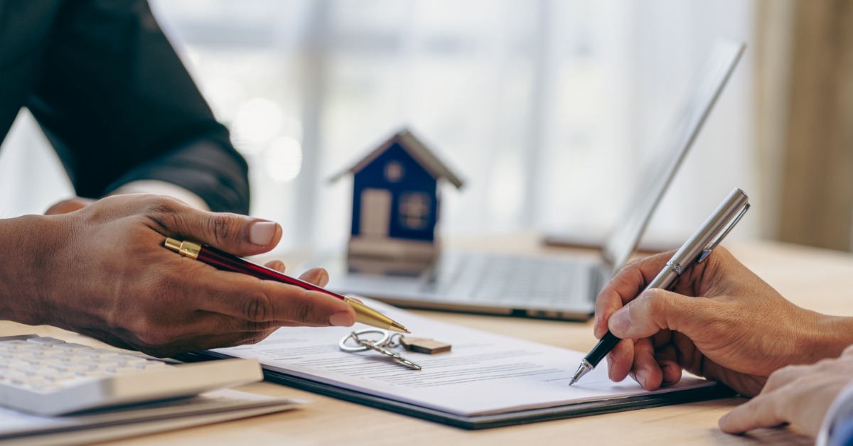 A person signing documents while another points with a pen. There are house keys, a laptop, and a model home nearby.