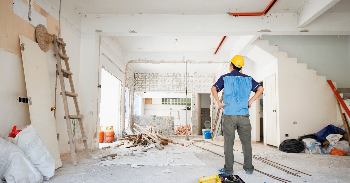 A person in a yellow hard hat standing in an unfinished room with ladders, debris, tools, and exposed walls.
