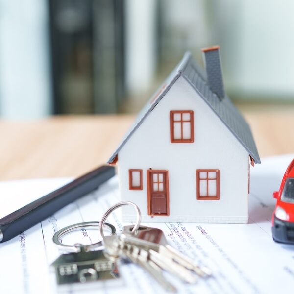 A small white model house, red toy car, keys, and black pen arranged on property paperwork on a table.