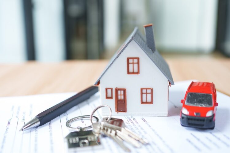 A small white model house, red toy car, keys, and black pen arranged on property paperwork on a table.