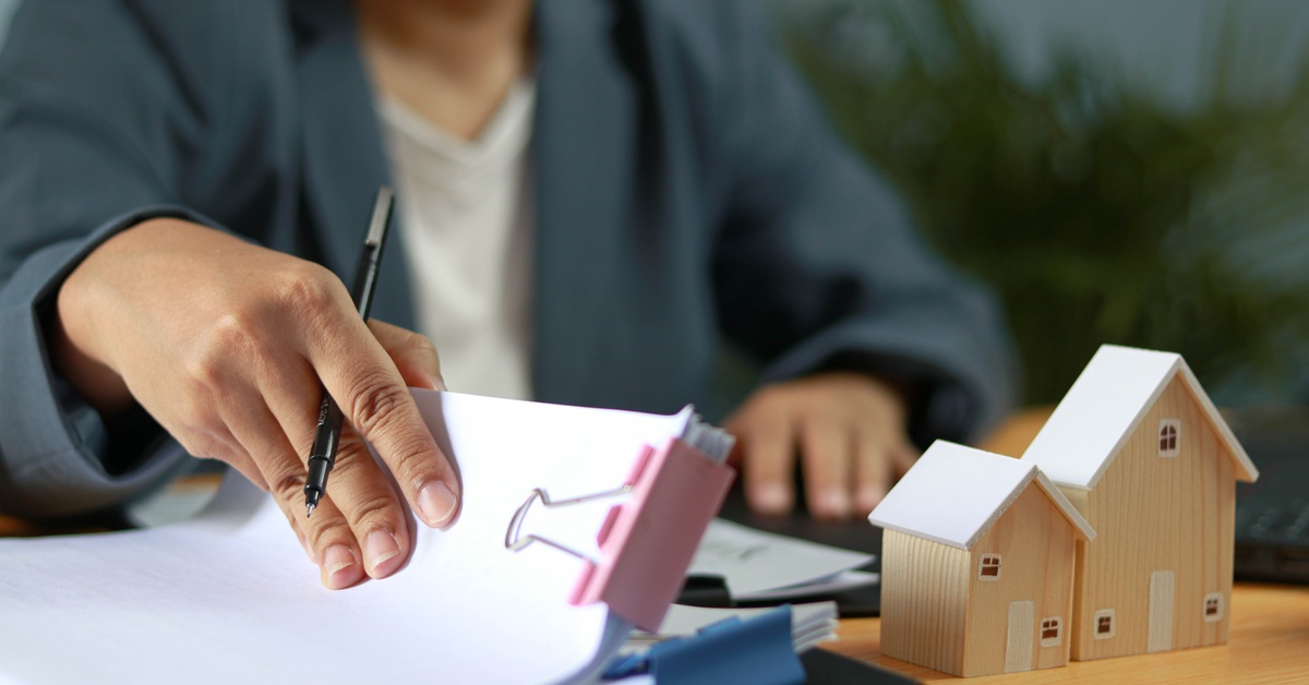 A person holding a pen over documents beside wooden model houses on a desk with folders and stacked papers.