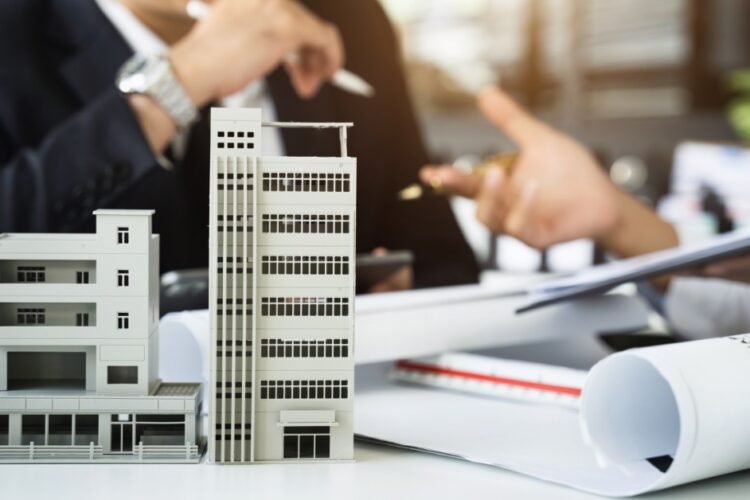 Miniature condominium models on a desk near a person holding a pen above a contract form on a clipboard.