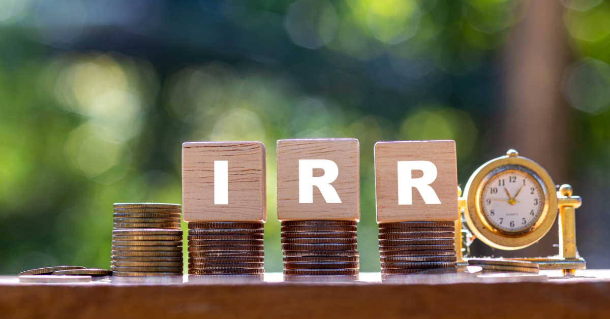 Stacks of coins topped with wooden IRR blocks beside a small gold clock against a blurred green background.