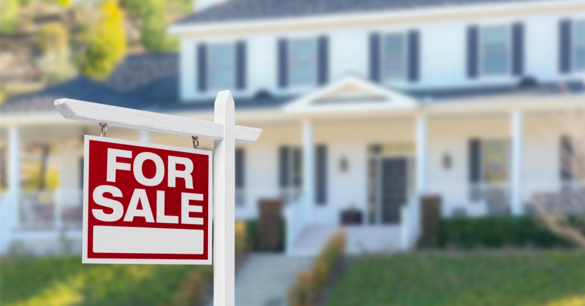 A red and white for sale sign standing on a lawn in front of a large blurred white house with dark shutters.