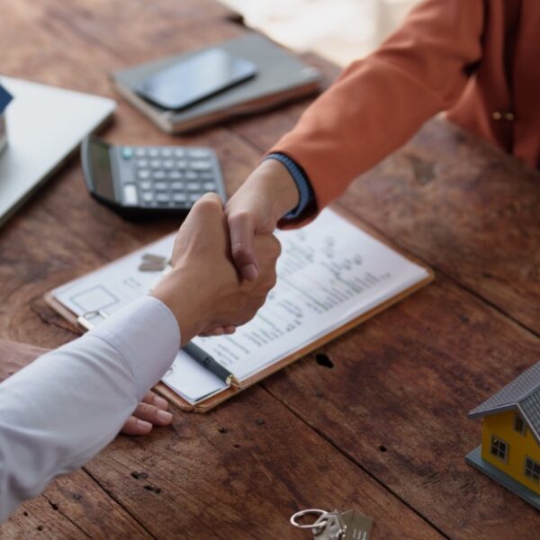 Two people shaking hands over a wooden table with house models, keys, a calculator, and paperwork on a clipboard.