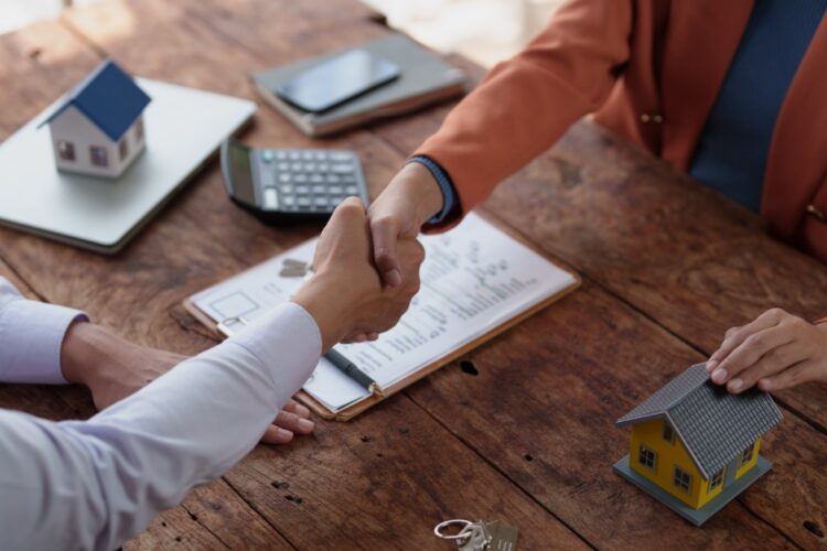 Two people shaking hands over a wooden table with house models, keys, a calculator, and paperwork on a clipboard.