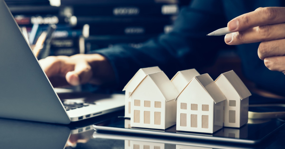 A person arranging three wooden house models on a table with upward arrows symbolizing rising property values.