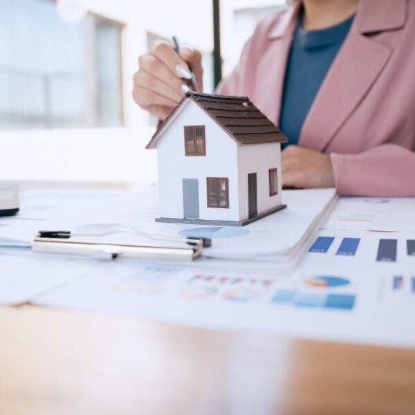 A woman reviewing financial charts at a desk with a small model house, laptop, calculator, and documents.
