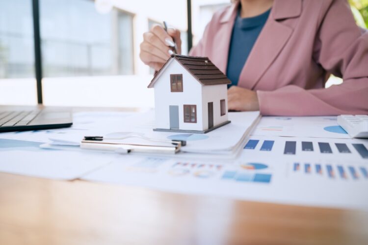 A woman reviewing financial charts at a desk with a small model house, laptop, calculator, and documents.