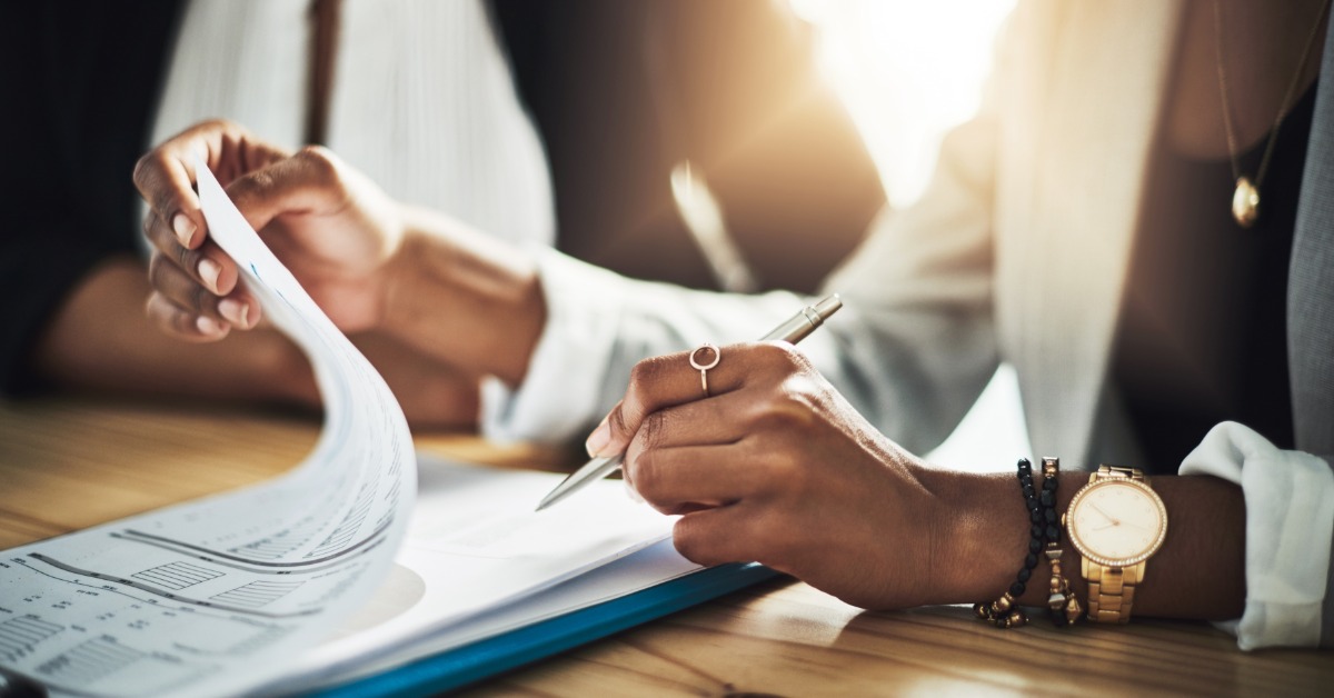 A close-up of two people reviewing paperwork at a wooden table, with one holding a pen and turning a page on a clipboard.