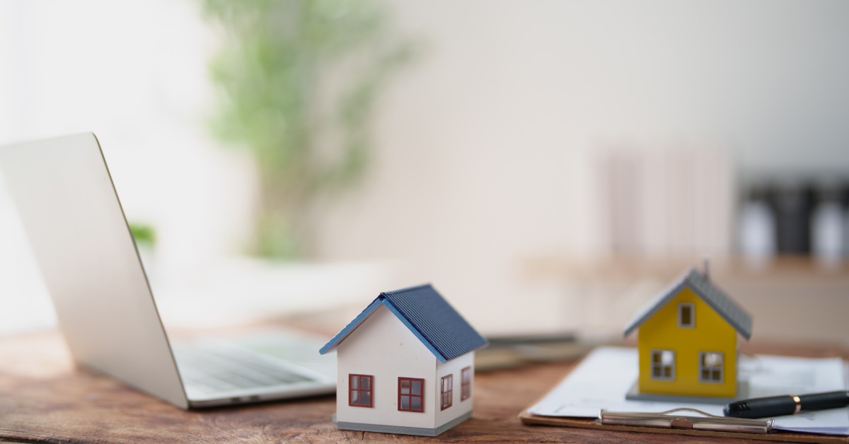 Two model houses set up on a wooden desk beside an open laptop, a clipboard with papers, and a black pen.