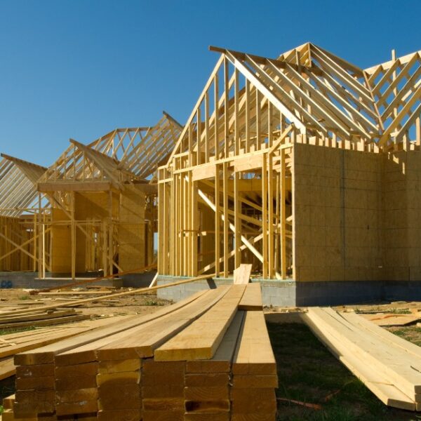A large house under construction with exposed wooden framing, roof trusses, and stacks of lumber outside.