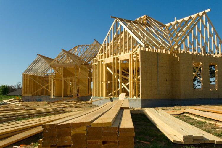 A large house under construction with exposed wooden framing, roof trusses, and stacks of lumber outside.