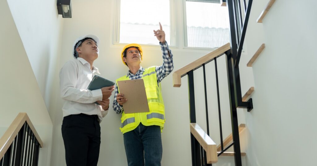 Two professionals wearing hard hats and holding clipboards while inspecting a stairwell. One is pointing upward.