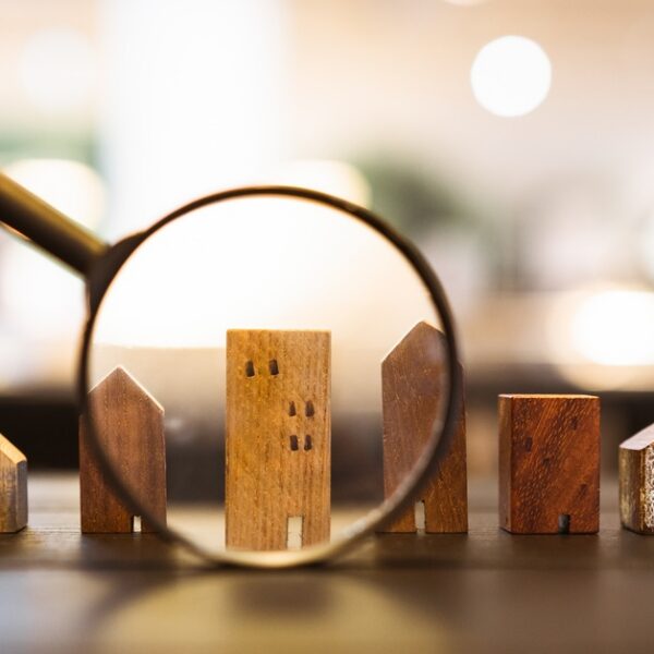 A hand holding a magnifying glass over small wooden house and building models arranged in a row on a table.
