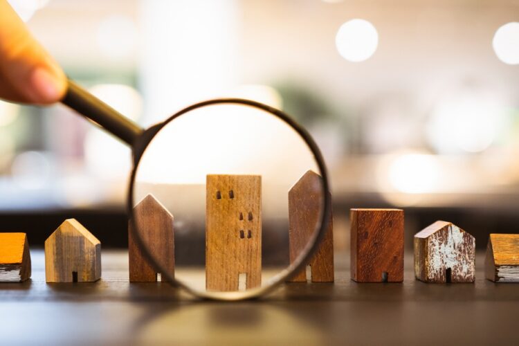 A hand holding a magnifying glass over small wooden house and building models arranged in a row on a table.
