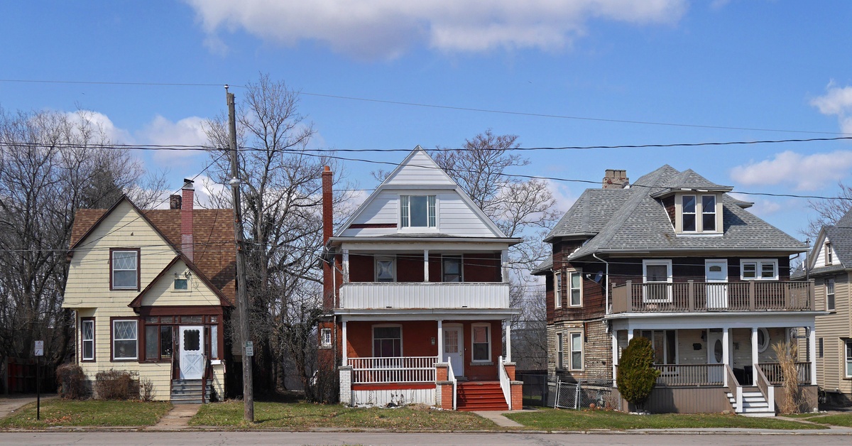 Three older two-story houses with front porches of different sizes standing side by side along a residential street.
