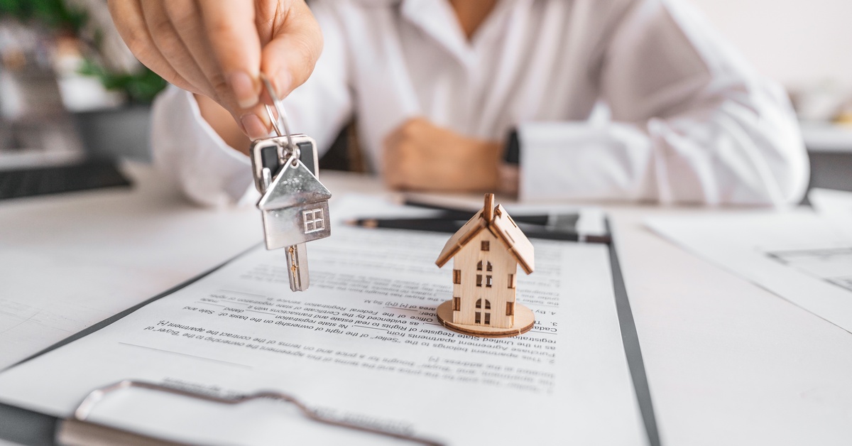 A close-up of a person holding a key with a house-shaped keychain above a property contract and a small wooden model home.