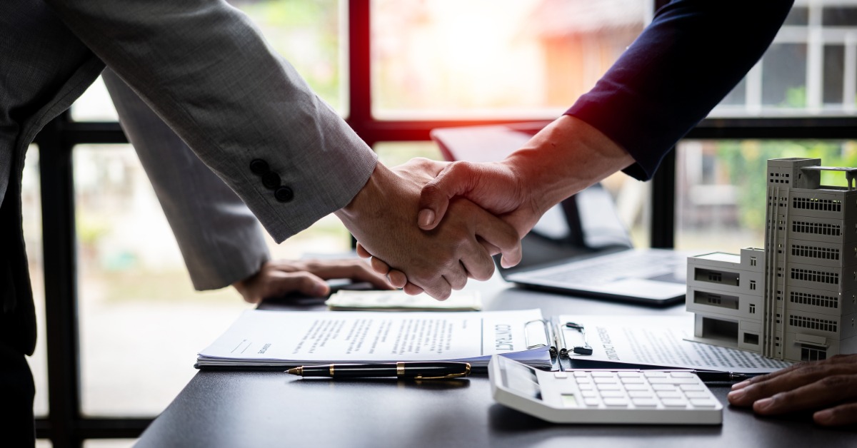 Two businesspeople shaking hands over a desk with loan documents, a calculator, a laptop, and building models.