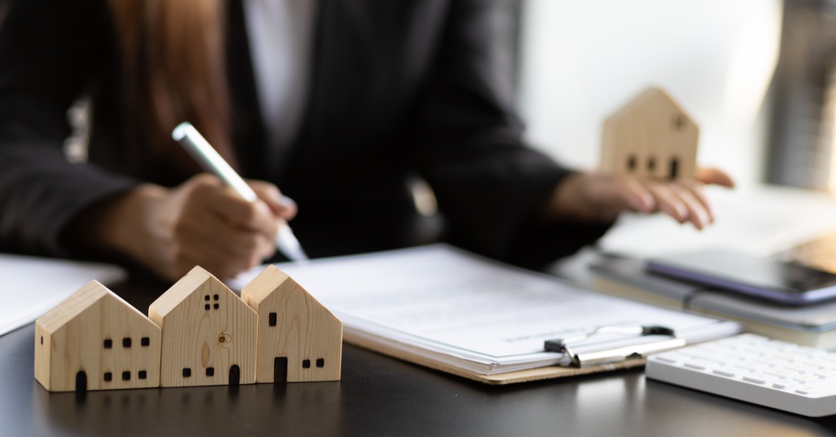 A businessperson writing on documents at a desk with wooden model houses, a calculator, and a laptop nearby.