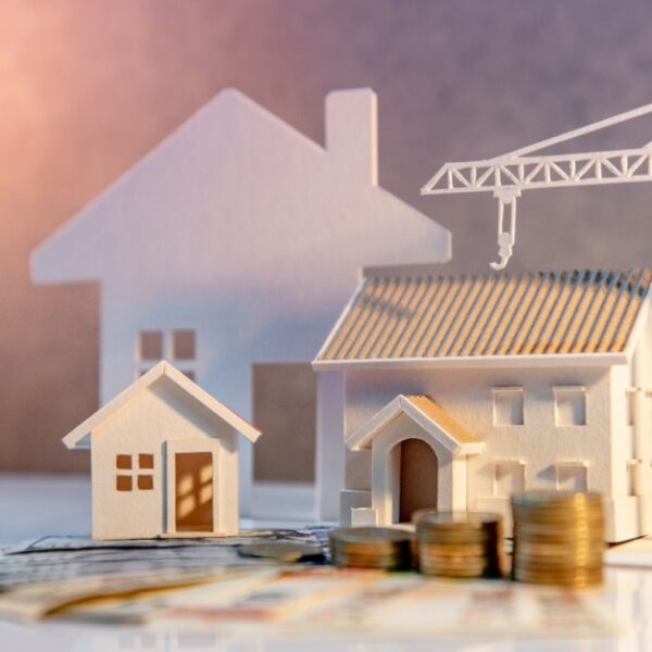 Miniature wooden houses, a white construction crane, stacked coins, and paper money representing home construction costs.