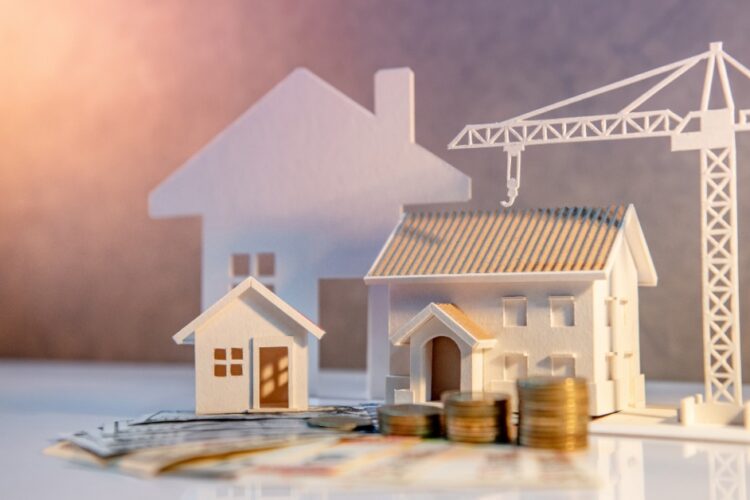 Miniature wooden houses, a white construction crane, stacked coins, and paper money representing home construction costs.