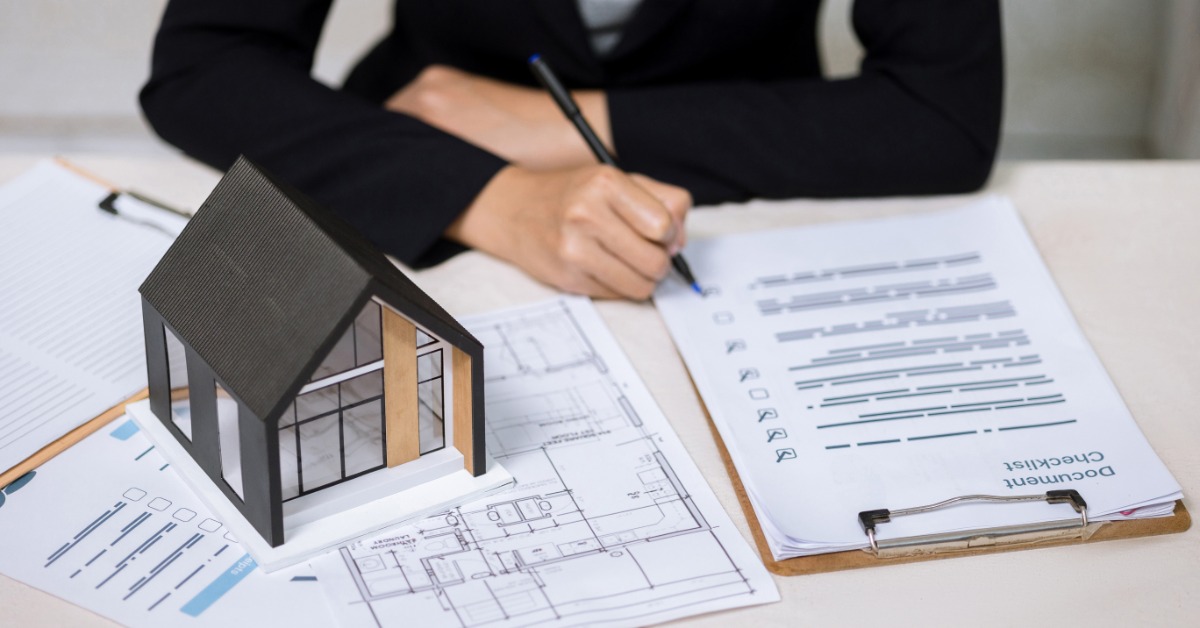 A person filling out a document checklist beside a miniature house model, architectural blueprints, and paperwork.